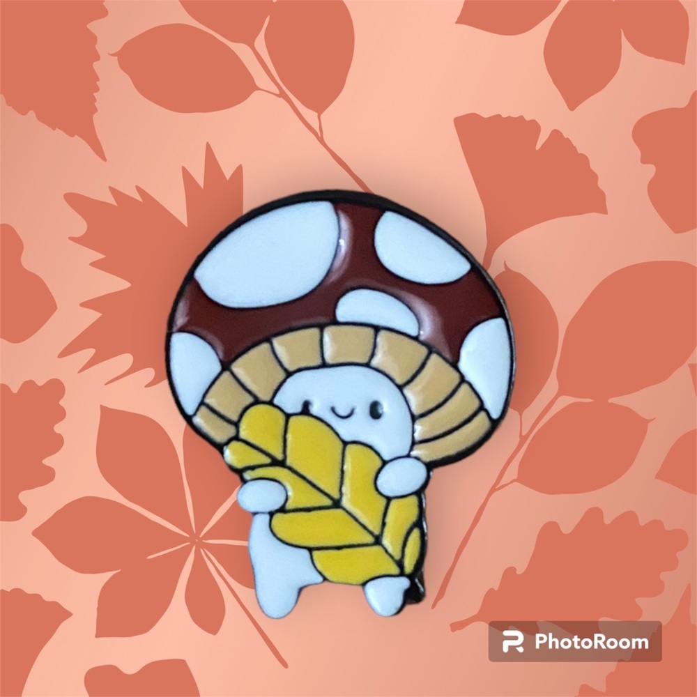 Red and white mushroom holding a leaf enamel pin